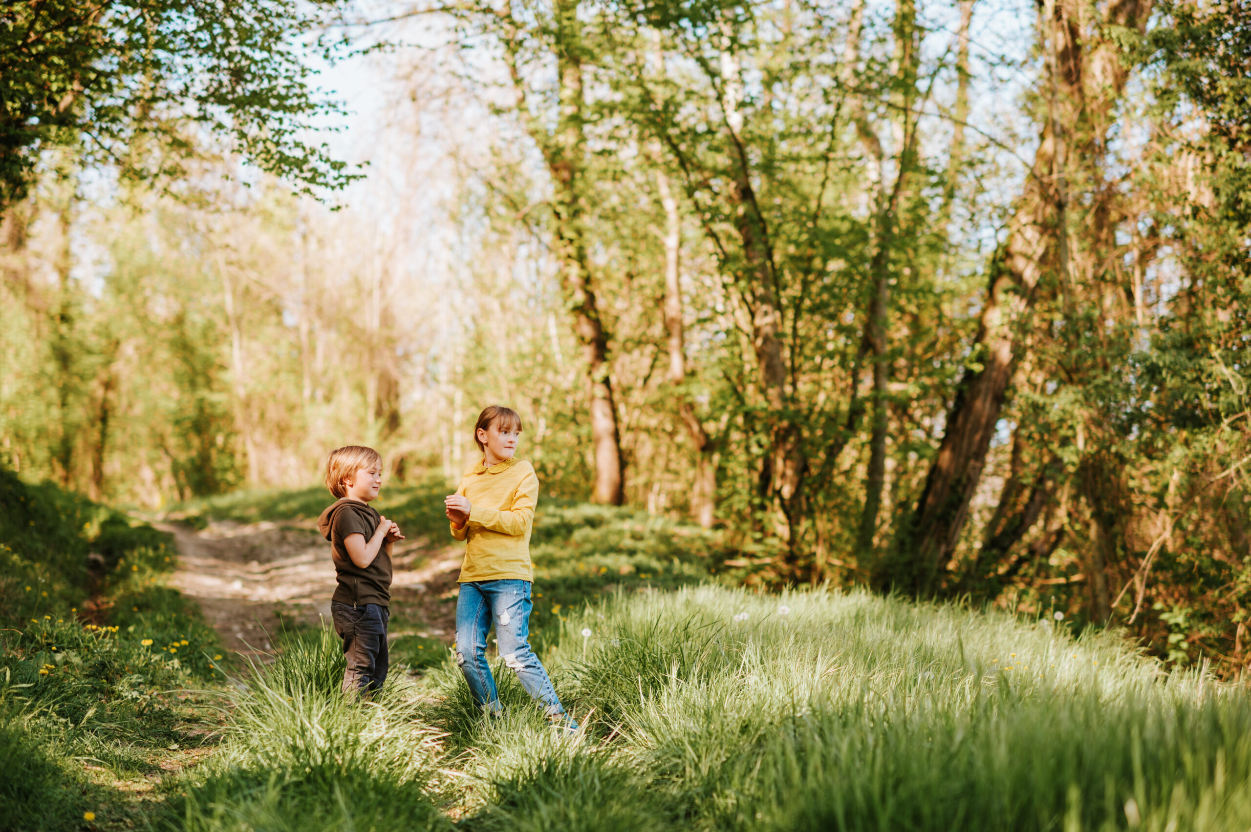 Two children, a boy and a girl, play together in a wooded outdoor setting, exploring among trees and brush.
